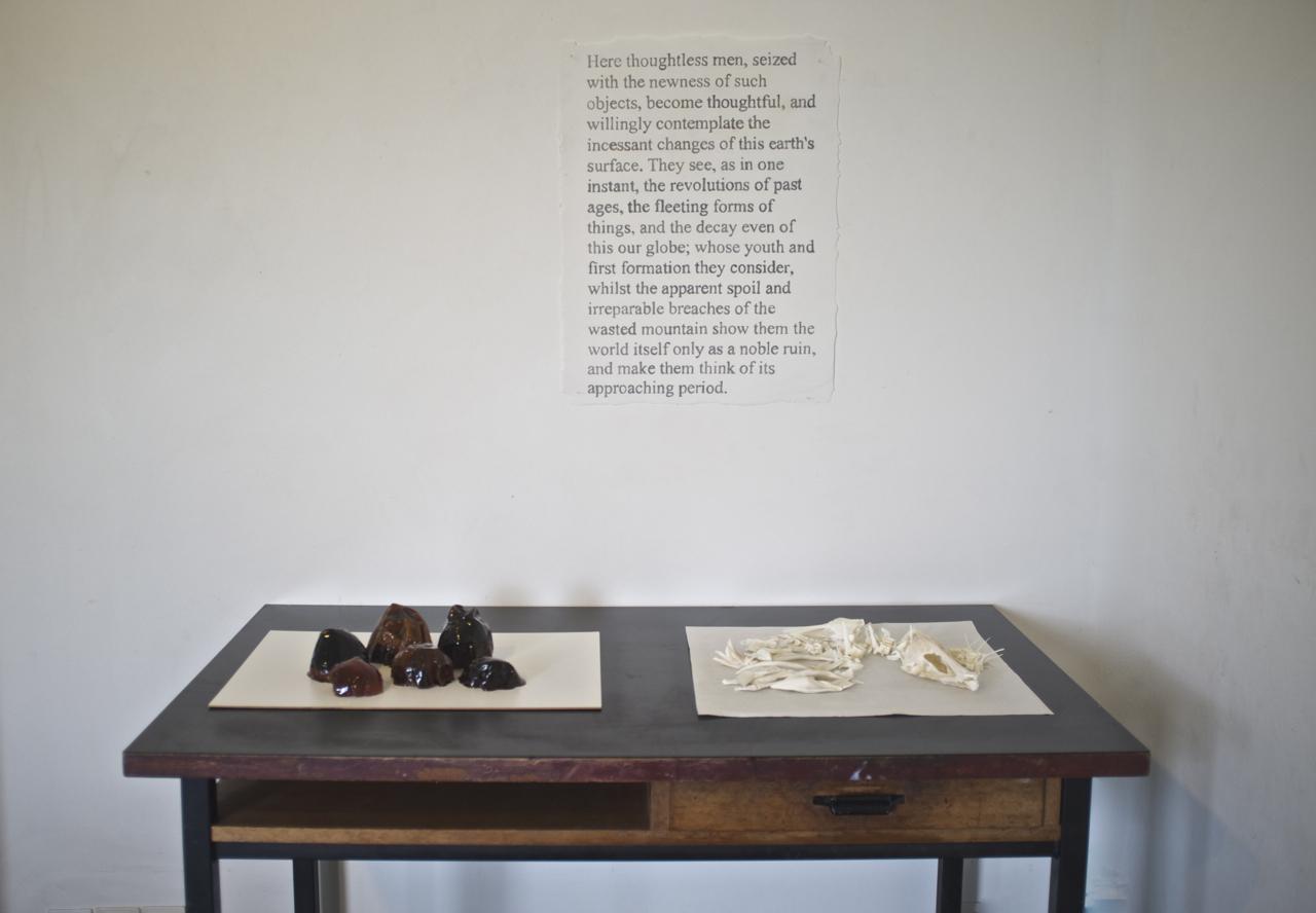 A photograph showing an artwork comprising a table with objects arranged on it, and above it a text