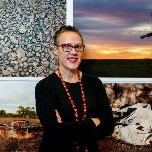 Brenda L Croft, who is standing in front of four artworks. She has her arms crossed and is smiling.