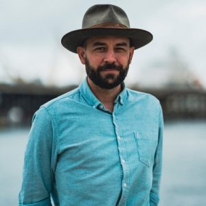 Clint Bracknell, who is wearing a hat and standing in front of some water.