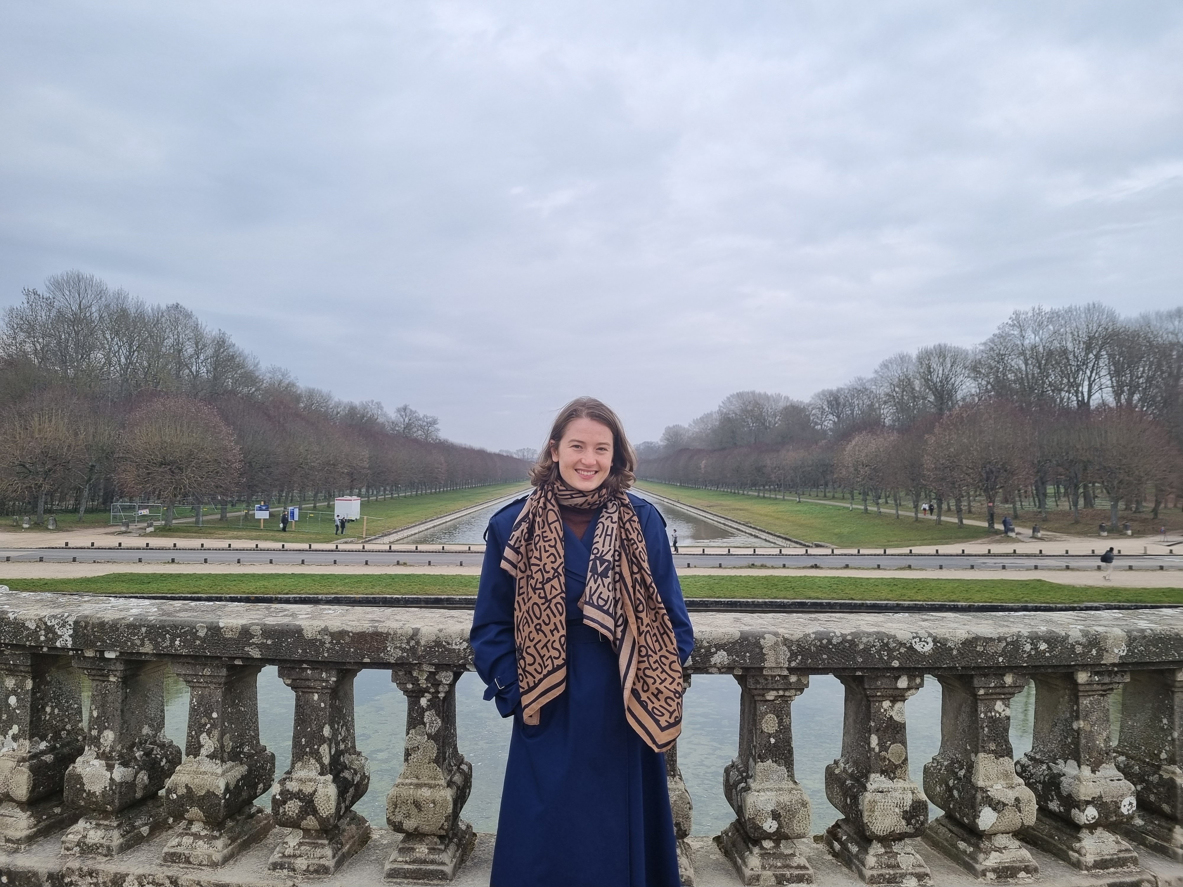 Imogen King stands in front of a low stone balustrade, with the forest at Fontainebleu behind her