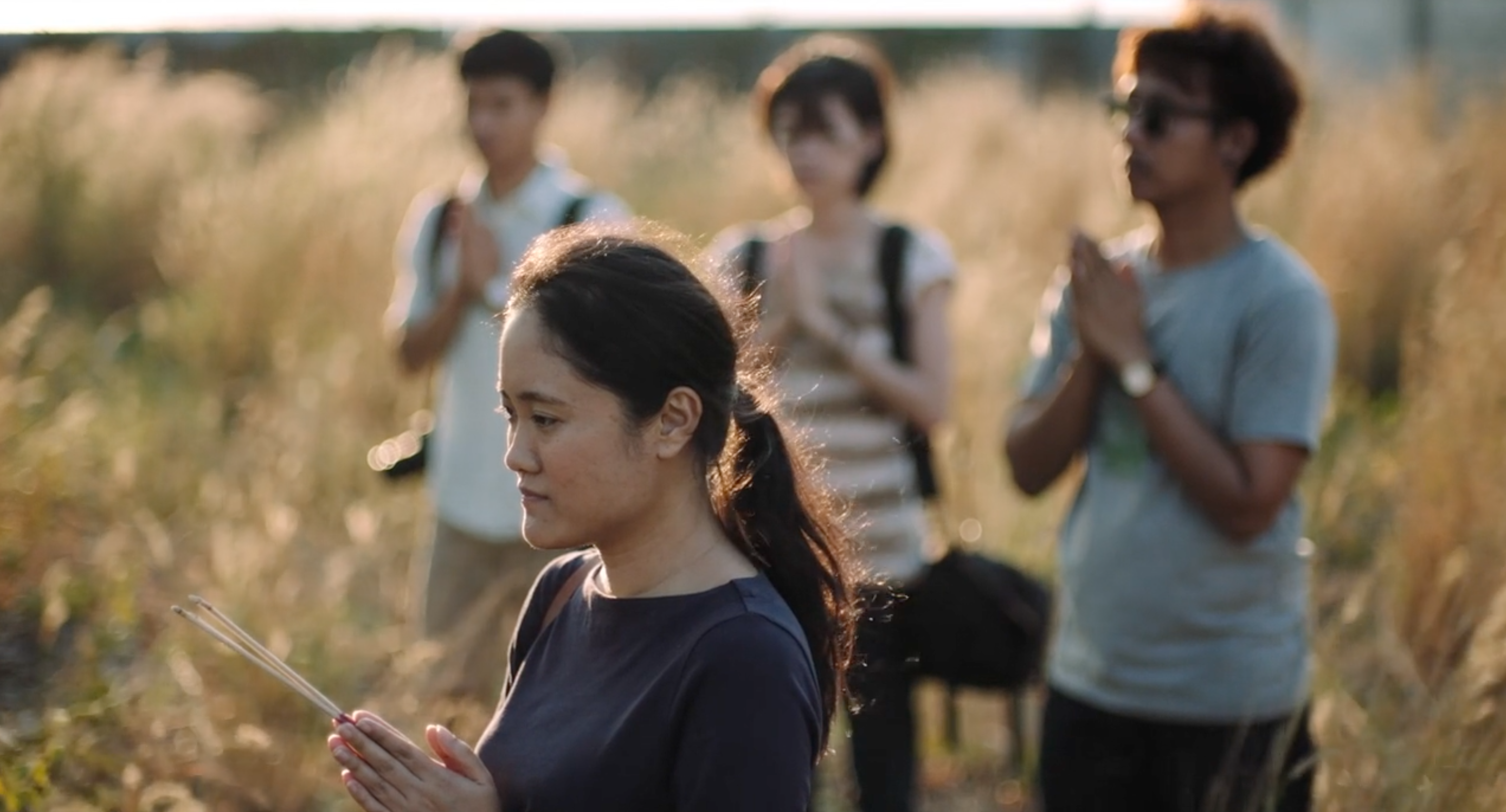 A still from the 2016 film "By the Time It Gets Dark", directed by Anocha Suwichakornpong, which shows four people standing in a field, with holds folded in prayer.