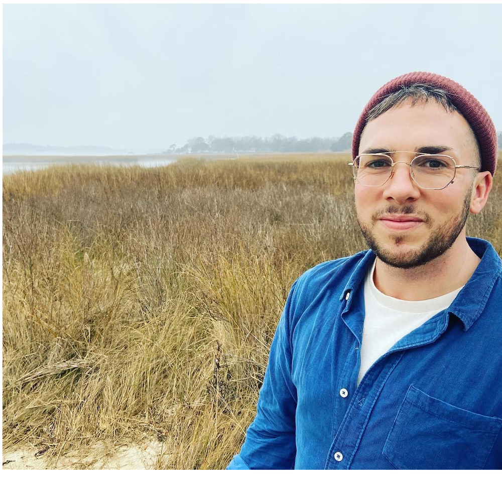 A white trans man with salt and pepper hair peeking out from beneath a mauve beanie and a trimmed beard stands amidst a salt marsh, wearing wire-rimmed glasses and a blue corduroy shirt.